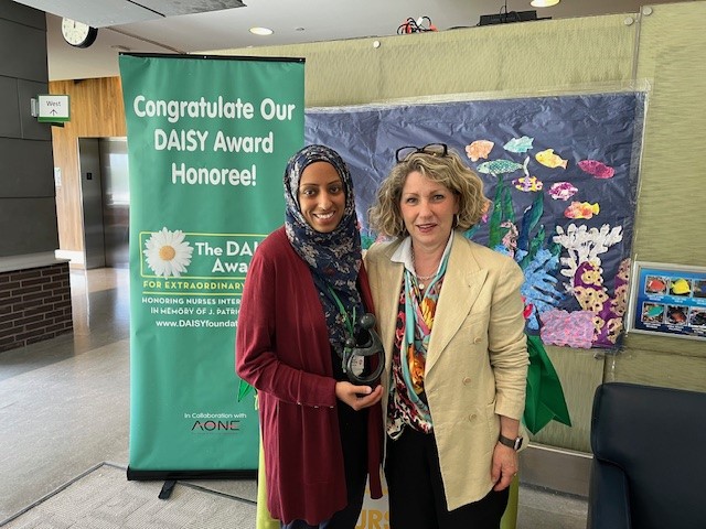 Two adults standing in front of a banner with the words "Congratulate our Daisy Award honoree!"