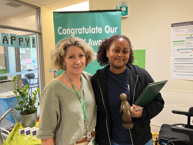 Two adults standing in front of a banner with the words "Congratulate our Daisy Award honoree!"
