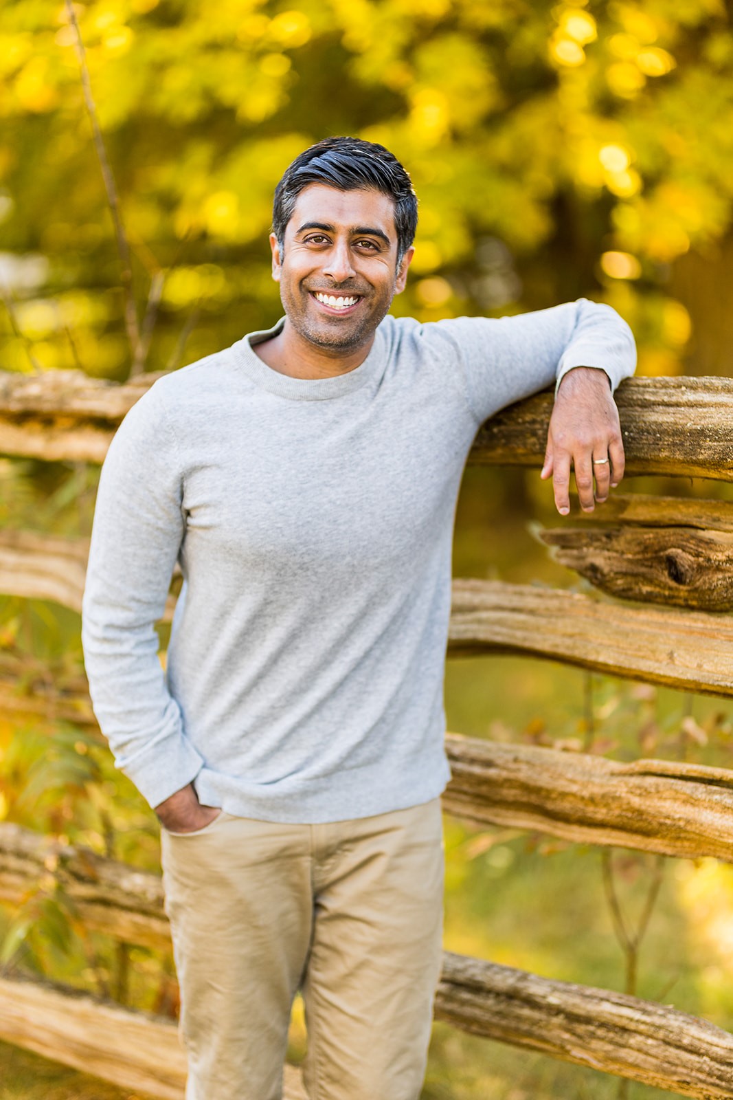 Dr Imran Khamis smiling and leaning on a wooden fence outdoors, wearing a light grey sweater and beige pants, with autumn foliage in the background.