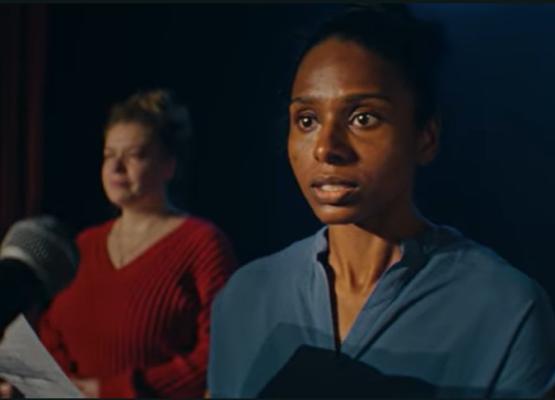 Woman in blue shirt stands in front of microphone while another woman is in the background