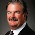 Man with brown hair and a moustache wearing a black blazer and striped tie