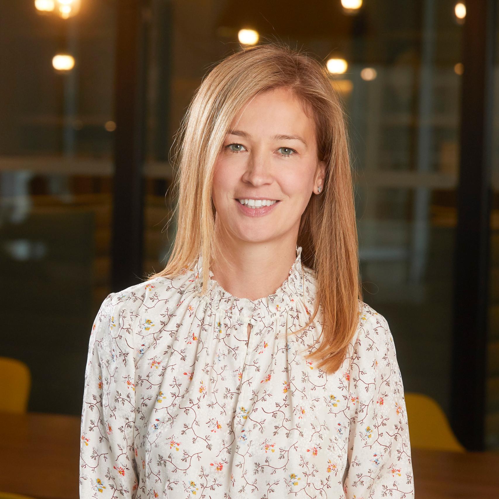 Woman with long blonde hair smiling. She is wearing a white floral blouse.