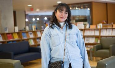 A young adult, who has a bandage on their neck, standing in a library, smiling