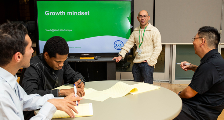 Presenter stands near screen with slide titled "Growth mindset", 3 youth sit at a table
