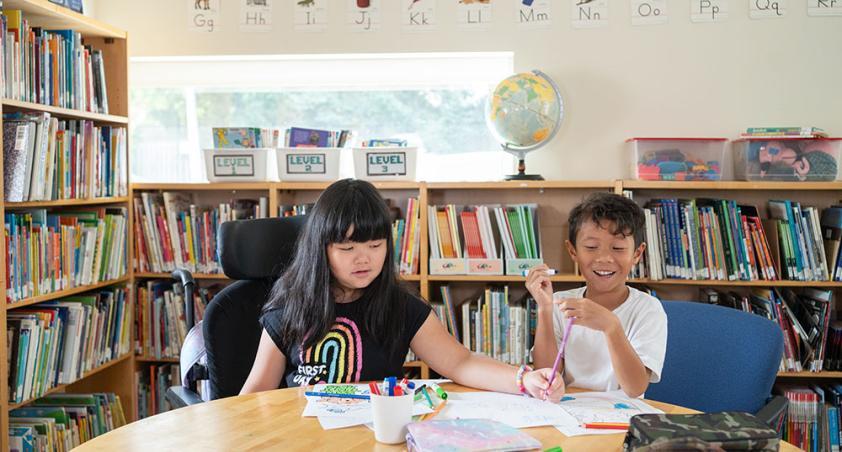 A child o wheelchair and another child to do some work on a desk, with a library setting at the back