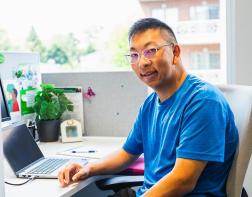 A young man sitting at a desk in front of a computer, looking towards the camera and smiling