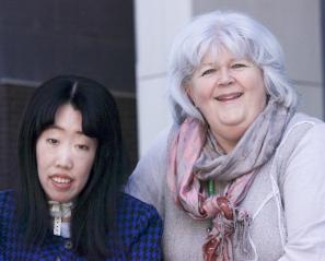 Woman with long black hair in navy sweater with a tracheotomy cork in her neck smiles with woman with grey hair wearing a pink and purple scarf