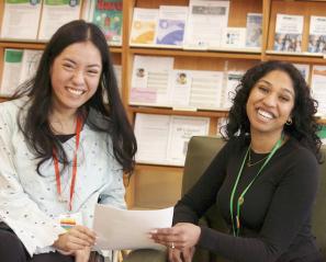 Two women with dark hair hold papers with shelves behind them
