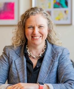 Julia Hanigsberg smiling and sitting in her office. She is wearing a blue blazer and her green Holland Bloorview staff lanyard. 