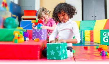 Young girl with dark hair sitting in a playroom with brightly coloured toys all around her. 