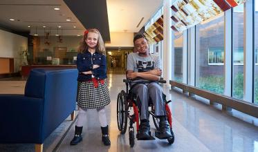 A child standing by the side of another child who is on a wheelchair