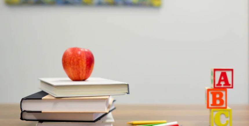 A small stack of books on a table with a red apple on top. A few coloured pencil crayons and three ABC blocks are stacked beside the books.