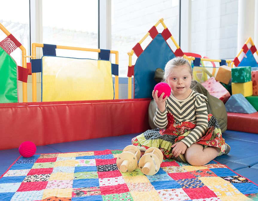 Little girl holding a red ball sitting on a quilt in a playroom