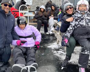 Teen in purple jacket on sledge holds hand of man beside girl in skates sitting on woman's lap