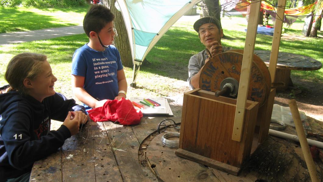 Two campers at table work with wire and talk to artist