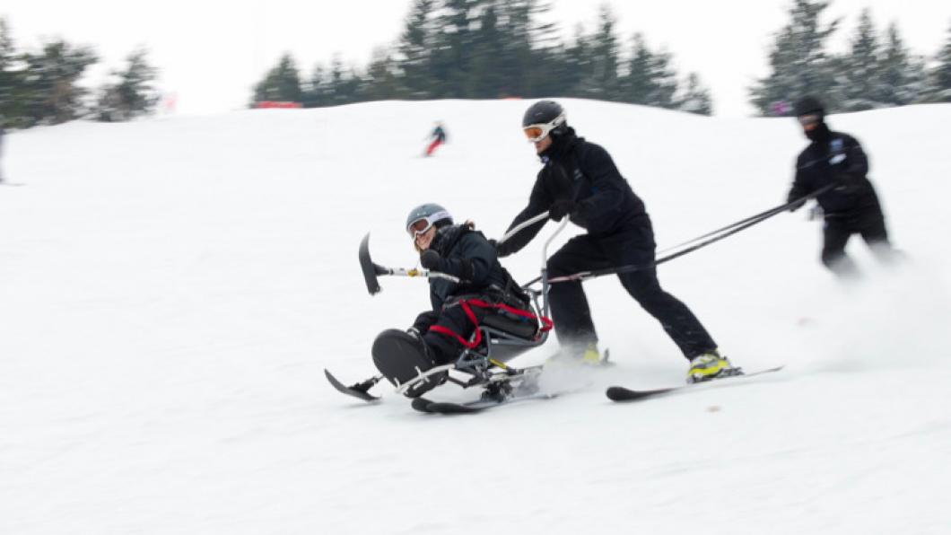 Woman descends hill on sit ski tethered to two volunteers