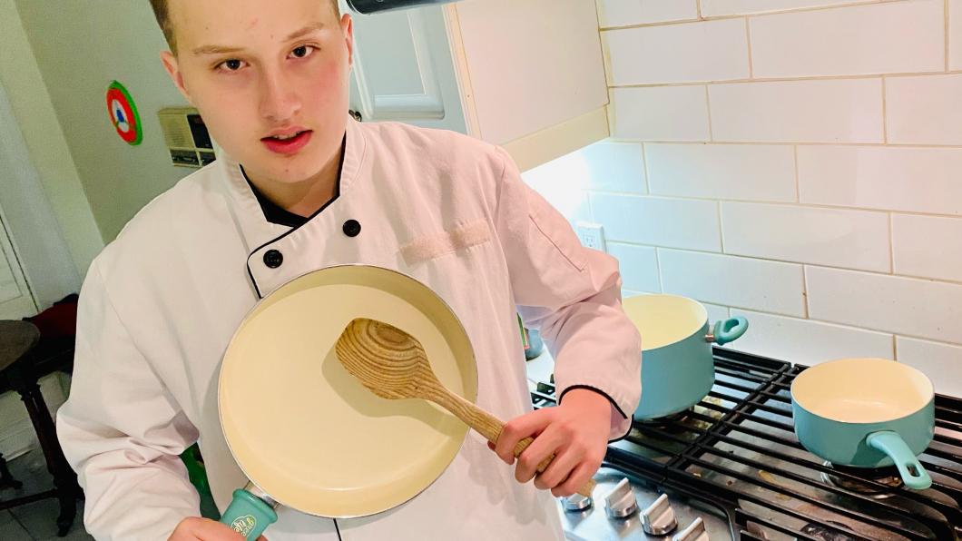 Mick in a chef's jacket holding up a pan and spoon in a kitchen.